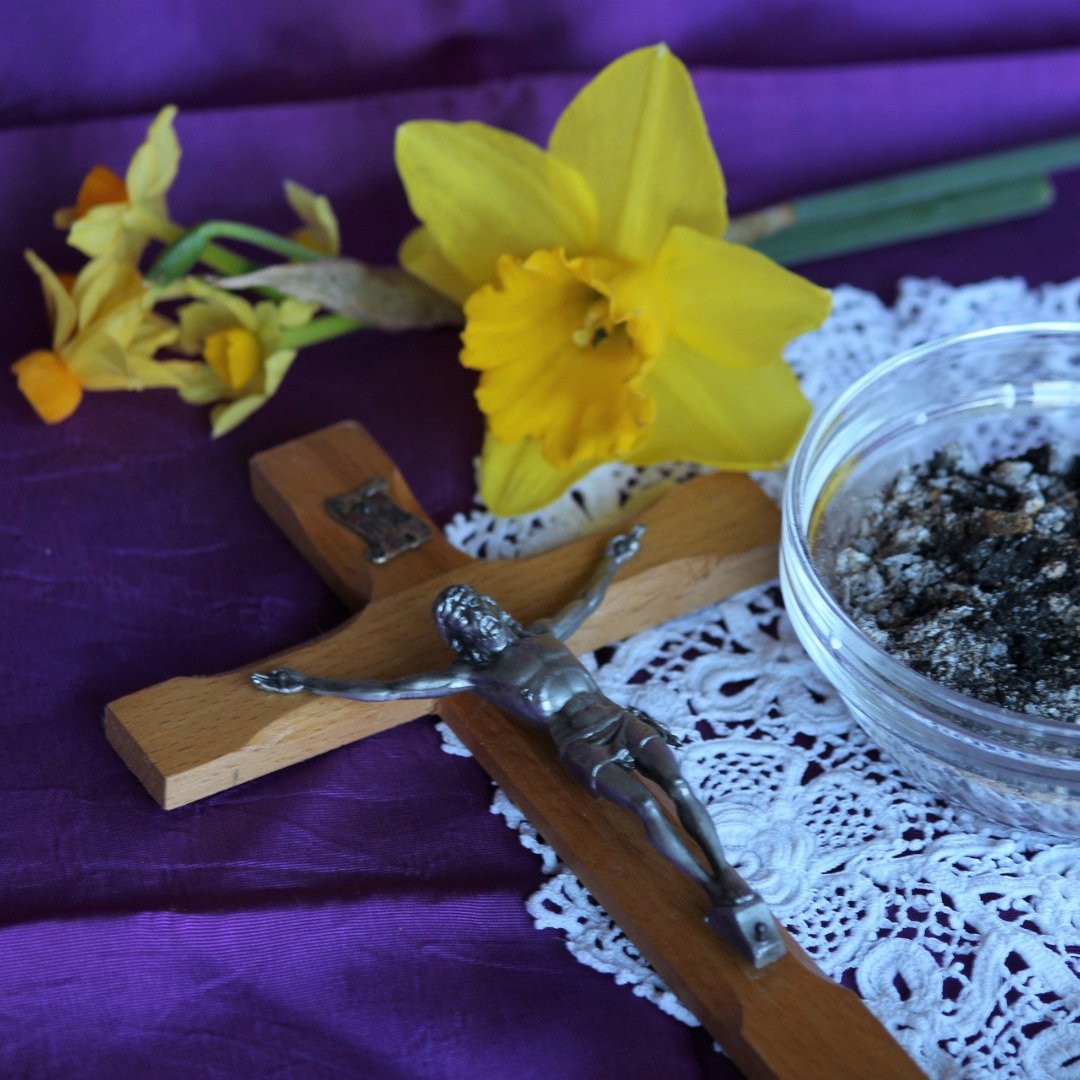 crucifix, daffodil, and bowl of ashes on purple tablecloth covered in white lace