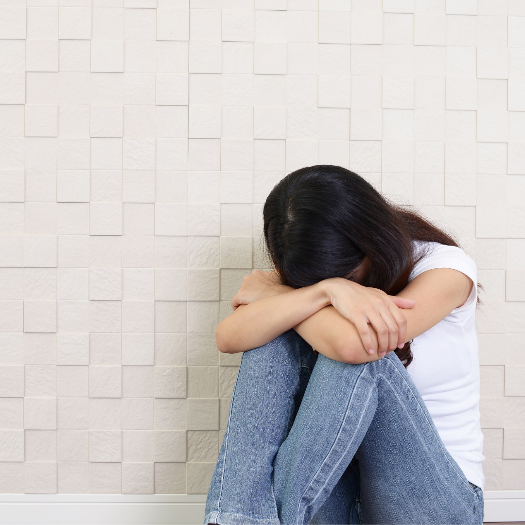 Dark-haired woman wearing jeans and white shirt sitting in front of white wall, hiding face