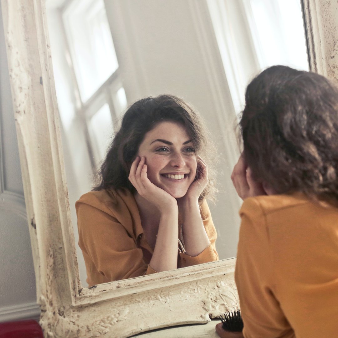 woman with long brown hair and orange blouse smiling at herself in vintage mirror