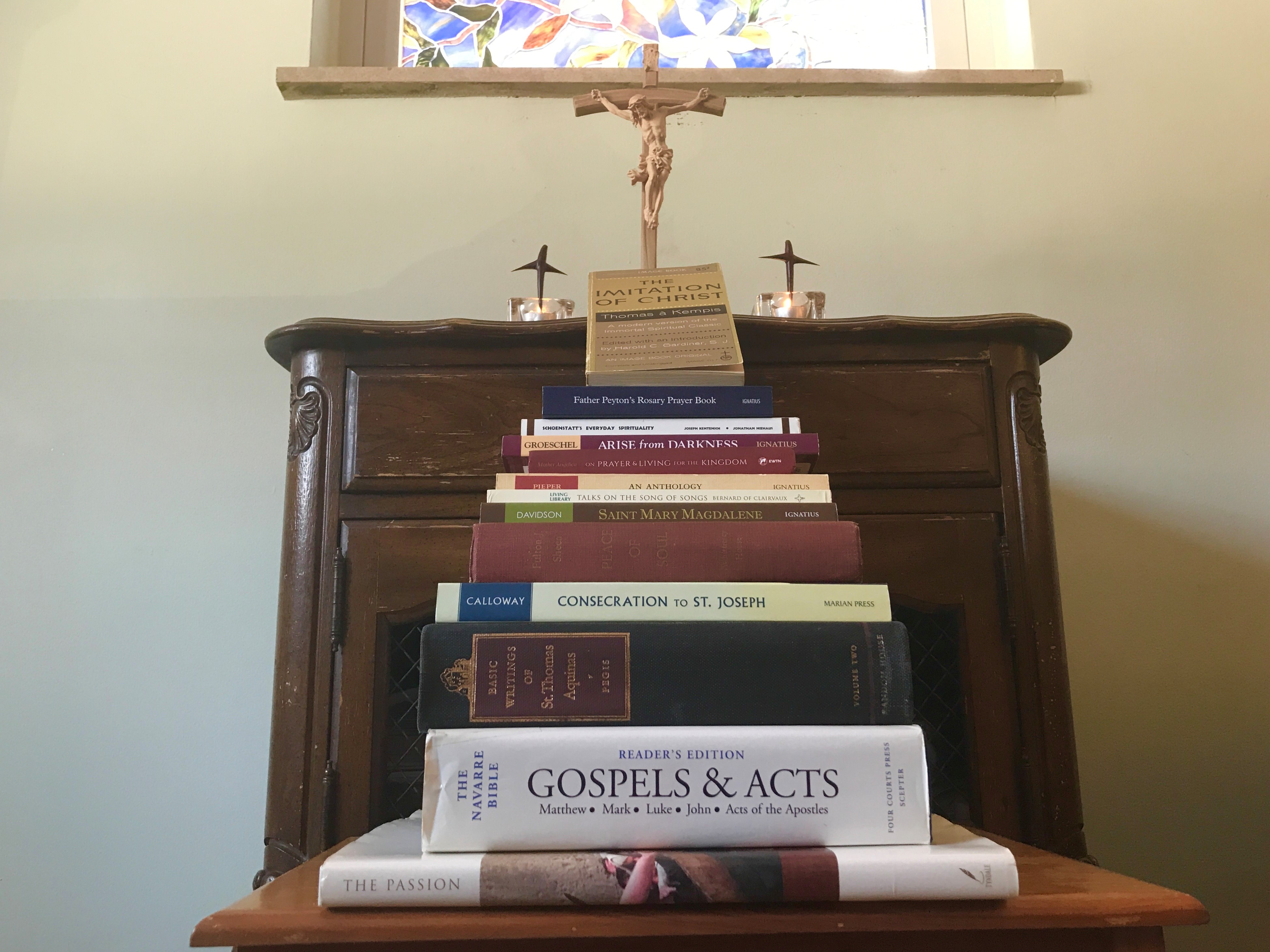 stack of spiritual books arranged stair-step-fashion with crucifix and candles on table at top.