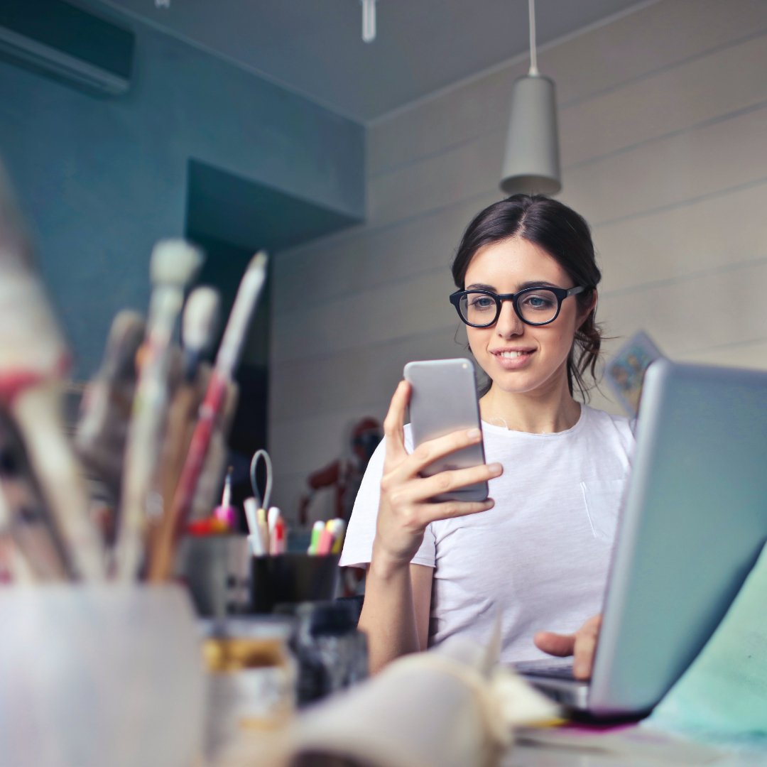 Woman with glasses and cell phone at desk covered in paint brushes, art supplies, computer