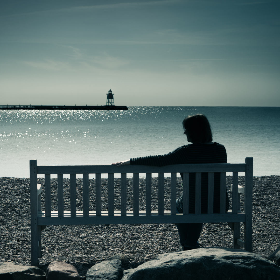 woman sitting alone on bench near shore, looking at lighthouse, stretching out her arm