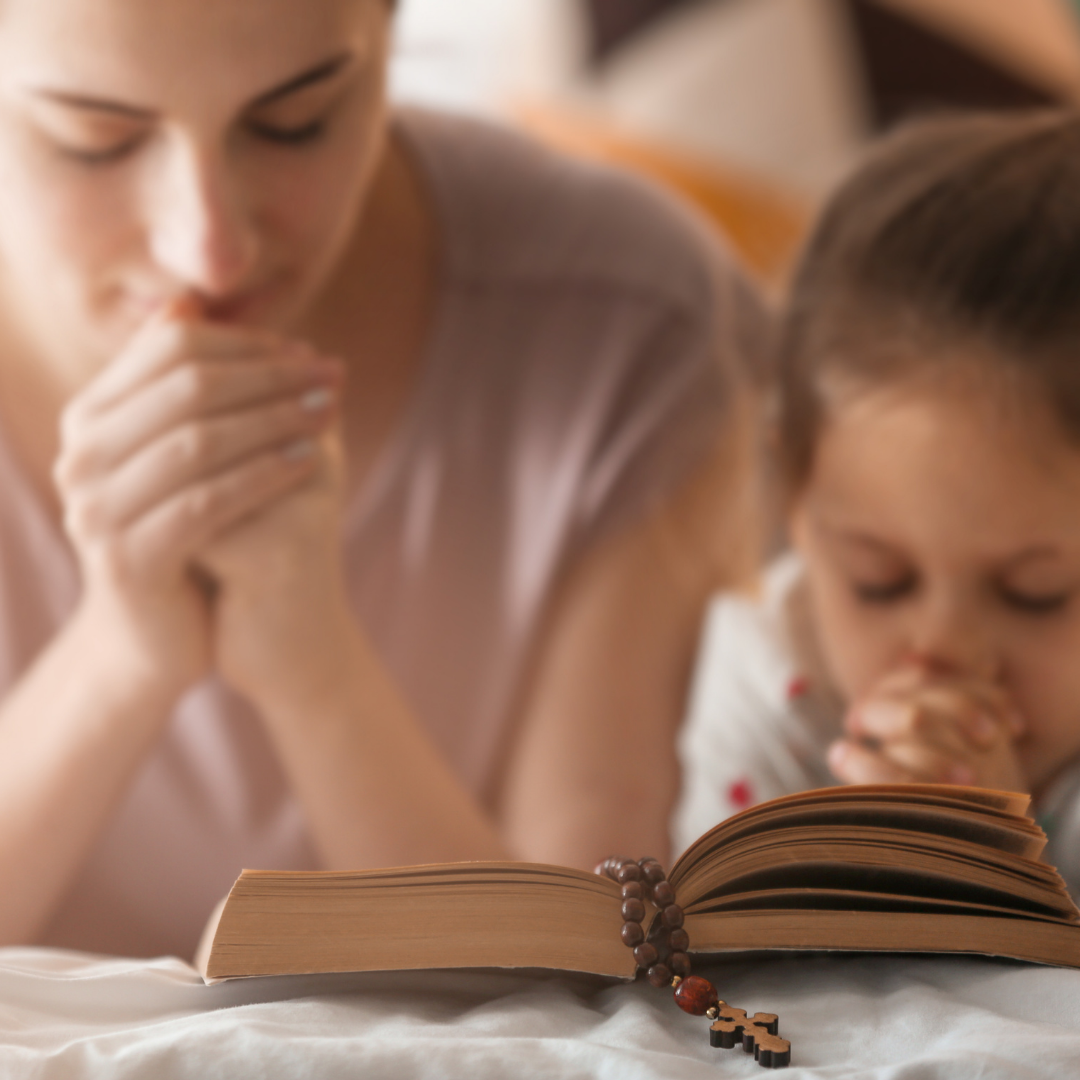 mom and little girl praying with open Bible and Rosary in front of them