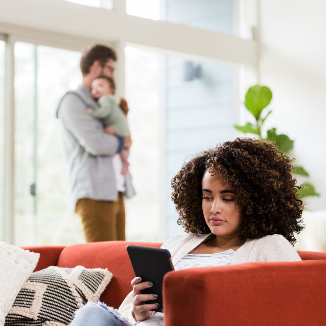 wife on phone with husband in the background holding baby