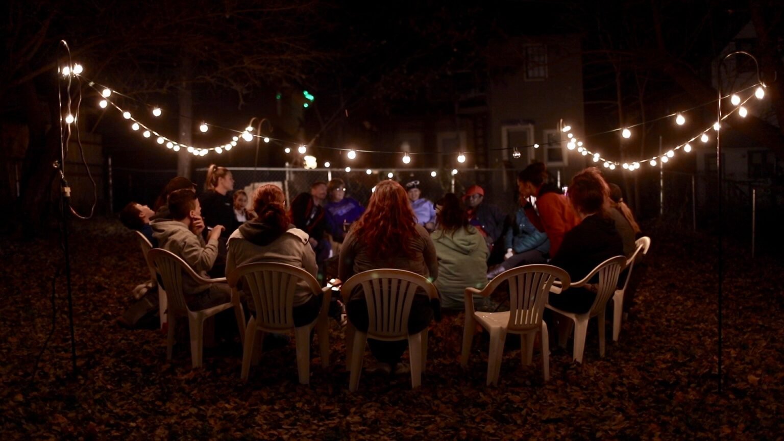 group of people sitting around a firepit in outdoor chairs with big white lights strung up around the yard