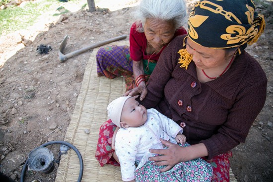 The Rana family lost their daughter Bhawana, age 5, when a courtyard collapsed on her in Kathmandu. Her surviving parents and infant sister have traveled to rural Taple to assist the elderly grandparents. Here, Bhawana's sister Bhumika (2 months) is tended to by her grandmother Modh Maya Rana, 71, in red, and mother Prem Kumari Rana. Jennifer Hardy/CRS