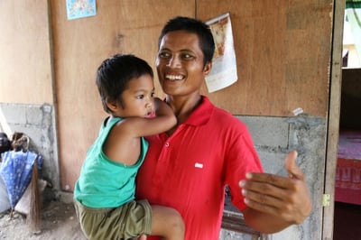 Ric De Veyra, age 30, had recently been elected Barangay chairperson but was still a month away from taking office when typhoon Haiyan / Yolanda devastated his community. When the sitting chairperson left for Manila immediately after the storm, it was up to Ric to lead the community in recovery. Here, Ric is pictured holding his 3-year-old son Joshua in front of his new CRS shelter. Jen Hardy/CRS